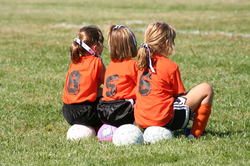 Three young girls waiting to be chosen to play football, sitting on footballs on the sidelines