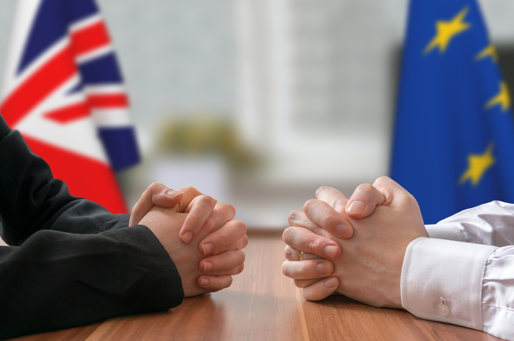 Two pairs of hands across a negotiating table with UK and EU flags behind